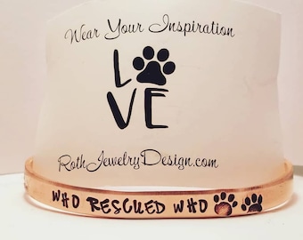 May include: A copper-coloured bracelet with the words "#D RESCUED #D" and two paw prints engraved on it. The bracelet is on a white background with a card that says "Wear Your Inspiration LOVE" with a paw print.