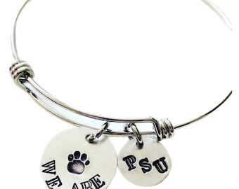 May include: A silver-colored bangle bracelet with two circular charms. One charm has a paw print and the words "WE ARE" stamped on it. The other charm has the letters "PSU" stamped on it. The bracelet is adjustable.