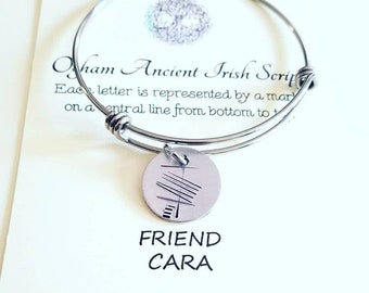 May include: A silver-toned bangle bracelet with a circular charm. The charm features an etched design and the words "FRIEND CARA". The bracelet is displayed on a card with text about Ancient Irish Script.