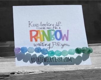 May include: A beaded bracelet with a rainbow of colors, resting in front of a white card. The card has the text "Keep looking up... there may be a rainbow waiting for you." and the Etsy shop name.