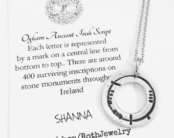 May include: A silver-colored pendant necklace with a circular design featuring Ogham script inscriptions. The necklace hangs on a delicate chain. A card behind the necklace describes the Ogham Ancient Irish Script and the name "SHANNA".