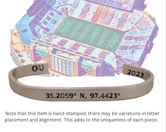 May include: A silver-colored cuff bracelet with the text "OU" and "2023" engraved on it. The bracelet also features the coordinates 35.2059° N, 97.4423°. The background shows a stadium illustration with the text "GAYLORD FAMILY - OKLAHOMA MEMORIAL".