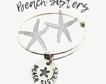 May include: Silver charm bracelet with a round charm that says "Beach Sisters" with a starfish in the centre. The bracelet has two starfish charms attached to it.