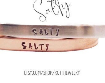 May include: Two stacked bracelets with the word "SALTY" engraved on each. The top bracelet is silver and the bottom bracelet is rose gold.