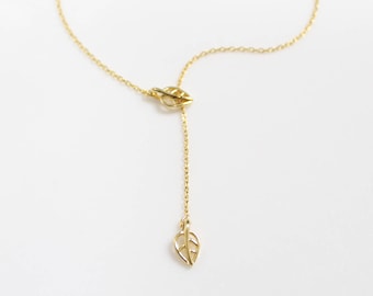 Könnte beinhalten: Eine goldfarbene Lariat-Halskette mit einer feinen Kette und zwei kleinen, durchbrochenen Blatt-Anhängern. Ein Blatt-Anhänger befindet sich an der zentralen Verbindungsstelle, während der zweite am Ende der vertikalen Kette herabhängt.