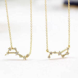 Può includere: Due collane d'oro con un designo di costellazione. Ogni collana presenta una catena delicata e un piccolo ciondolo di costellazione con piccole pietre trasparenti.