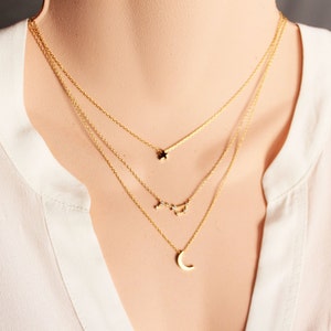 Può includere: Un set di collane dorate a strati con tre sottili catene. La catena superiore ha un piccolo ciondolo a stella, la catena centrale ha un design a costellazione e la catena inferiore ha un ciondolo a mezzaluna. Le collane sono mostrate contro una camicetta bianca.