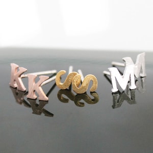 May include: Three pairs of initial earrings. The first pair is rose gold and features the letter K. The second pair is gold and features the letter S. The third pair is silver and features the letter M.