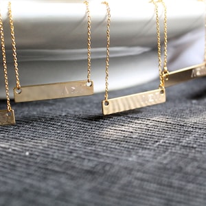 Op de afbeelding: Vijf gouden kettingen met delicate kettingen en gepersonaliseerde staafhangers. De hangers zijn rechthoekig en hebben een gehamerde textuur.