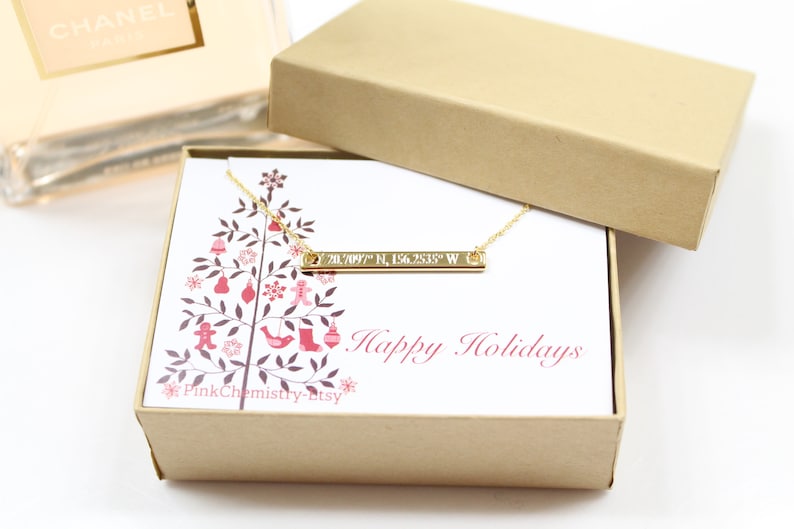 Puede incluir: Un collar de barra de oro con las coordenadas "70.7097&deg; N, 156.2535&deg; W" grabadas en &eacute;l. El collar est&aacute; en una peque&ntilde;a caja de regalo con un dise&ntilde;o de &aacute;rbol de Navidad y el texto "Felices Fiestas".