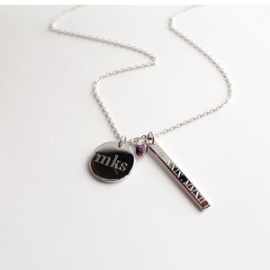May include: Silver necklace with a delicate chain. The necklace features a round pendant engraved with "mks", a rectangular bar pendant with Roman numerals, and small purple beads. The necklace is displayed on a white surface.