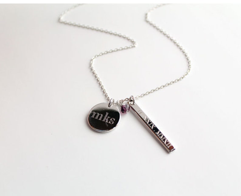 May include: Silver necklace with a delicate chain. The necklace features a round pendant engraved with "mks", a rectangular bar pendant with Roman numerals, and small purple beads. The necklace is displayed on a white surface.