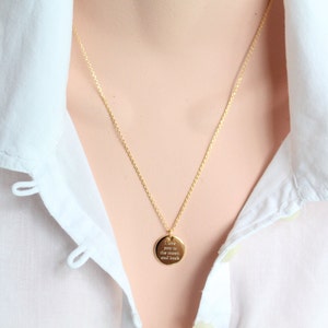 May include: A gold-colored necklace with a delicate chain and a round pendant. The pendant has the engraved text "I love you to the moon and back". The necklace is worn over a white collared shirt.