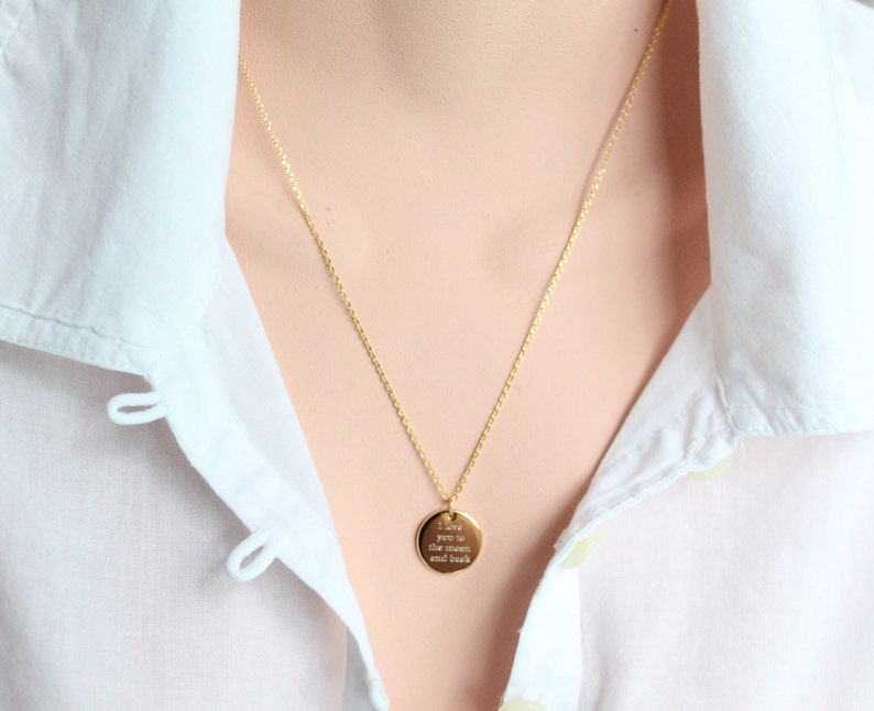 May include: A gold-colored necklace with a delicate chain and a round pendant. The pendant has the engraved text "I love you to the moon and back". The necklace is worn over a white collared shirt.