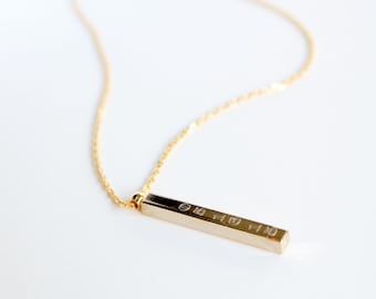 Op de afbeelding: Een gouden ketting met een lange, dunne, rechthoekige hanger. De hanger heeft gegraveerde tekst.