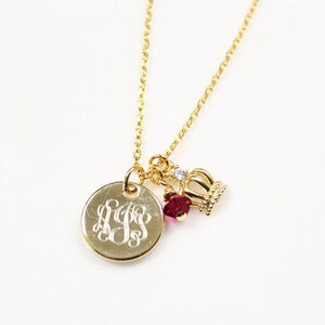May include: Gold-colored necklace with a round pendant featuring engraved initials. The necklace also includes a small crown charm, a red gem, and a small clear gem. The chain is delicate and the background is white.