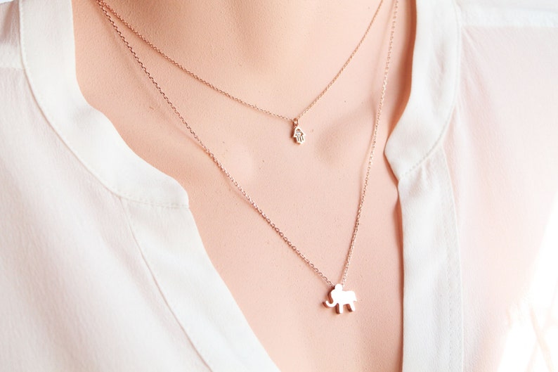 May include: Rose gold layered necklace with two pendants: a small Hamsa hand and an elephant. The necklaces are worn over a white blouse. The jewelry is delicate and elegant.