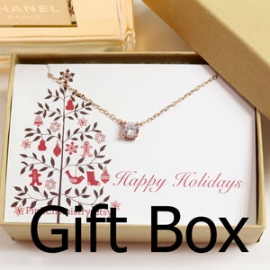 May include: A small gift box with a white background and a red and brown Christmas tree design. The box contains a delicate rose gold necklace with a clear cubic zirconia pendant. The box lid has the text "Happy Holidays" printed in red cursive font.