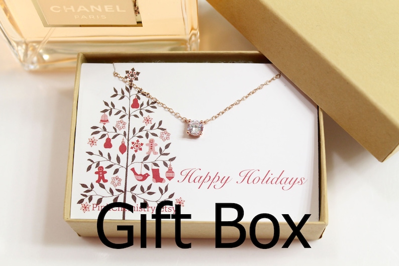 May include: A small gift box with a white background and a red and brown Christmas tree design. The box contains a delicate rose gold necklace with a clear cubic zirconia pendant. The box lid has the text "Happy Holidays" printed in red cursive font.