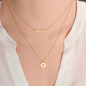 Puede incluir: Un collar de oro con dos cadenas. La cadena superior tiene la palabra "mommy". La cadena inferior tiene un peque&ntilde;o colgante redondo con el s&iacute;mbolo astrol&oacute;gico de Sagitario.