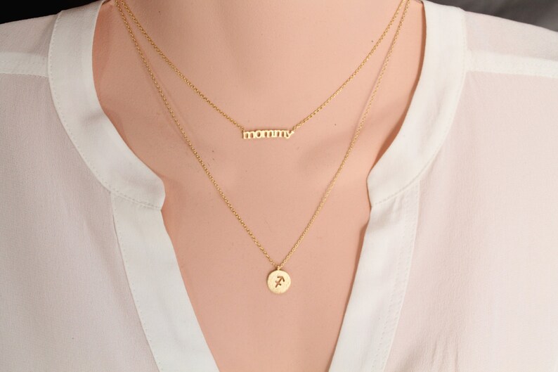 Puede incluir: Un collar de oro con dos cadenas. La cadena superior tiene la palabra "mommy". La cadena inferior tiene un peque&ntilde;o colgante redondo con el s&iacute;mbolo astrol&oacute;gico de Sagitario.