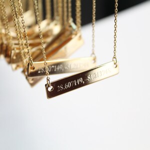 Op de afbeelding: Een gouden ketting met een rechthoekige hanger gegraveerd met de co&ouml;rdinaten "28.607149, -81.213748".