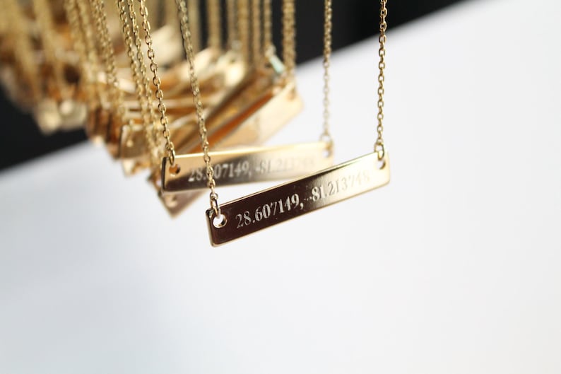 Op de afbeelding: Een gouden ketting met een rechthoekige hanger gegraveerd met de co&ouml;rdinaten "28.607149, -81.213748".