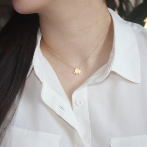 May include: A gold-colored elephant pendant necklace. The small charm is suspended from a delicate chain. The necklace is worn over a white collared shirt.