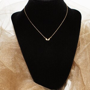Op de afbeelding: Een delicate gouden ketting met een klein, vleugelvormig hangertje. De ketting wordt getoond op een zwarte fluwelen standaard tegen een gouden, glinsterende achtergrond.