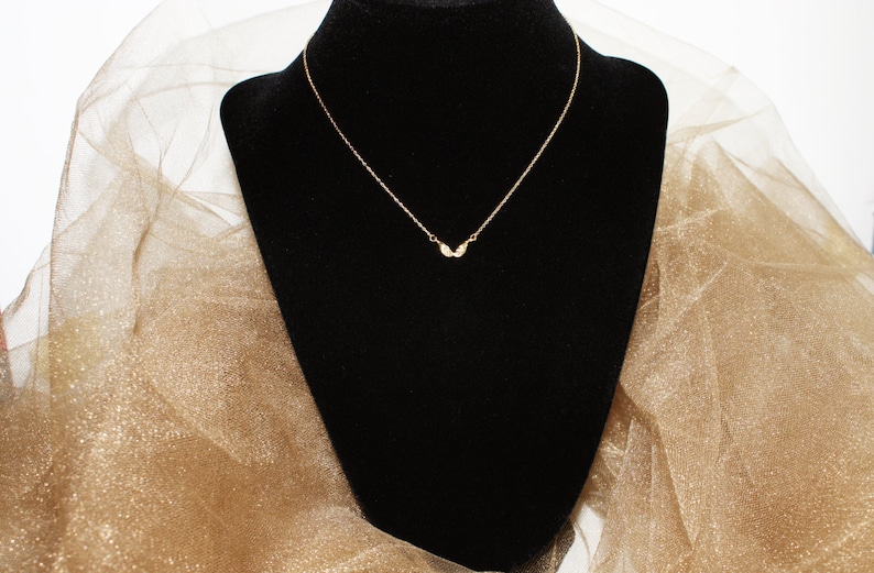 Op de afbeelding: Een delicate gouden ketting met een klein, vleugelvormig hangertje. De ketting wordt getoond op een zwarte fluwelen standaard tegen een gouden, glinsterende achtergrond.