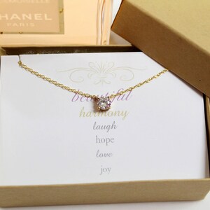 Puede incluir: Un collar de oro con un solo colgante de cristal transparente en una caja de regalo marr&oacute;n. La caja tiene una tarjeta blanca en el interior con las palabras "beautiful harmony laugh hope love joy".