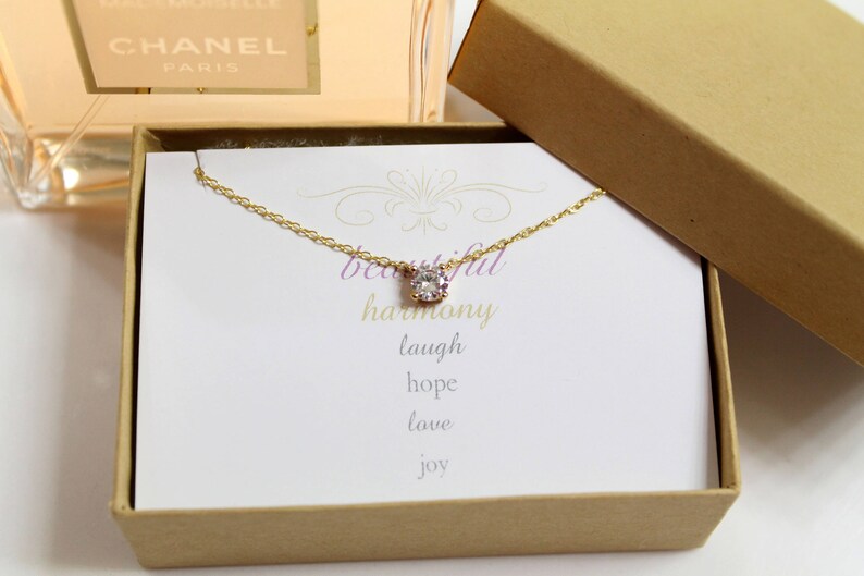 Puede incluir: Un collar de oro con un solo colgante de cristal transparente en una caja de regalo marr&oacute;n. La caja tiene una tarjeta blanca en el interior con las palabras "beautiful harmony laugh hope love joy".