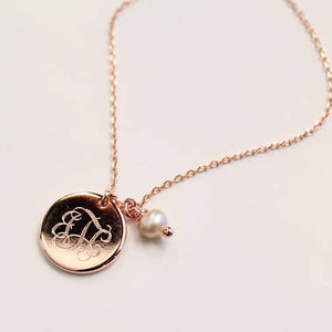 May include: Rose gold necklace featuring a round pendant with an engraved initial and a small pearl charm. The delicate chain and pendant have a warm, metallic sheen, creating an elegant and personalized accessory.