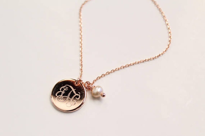 May include: Rose gold necklace featuring a round pendant with an engraved initial and a small pearl charm. The delicate chain and pendant have a warm, metallic sheen, creating an elegant and personalized accessory.