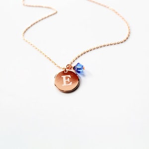May include: A rose gold necklace with a delicate chain. The necklace features a circular pendant engraved with the letter "E" and a small blue crystal charm. The pendant and charm are suspended from the chain.