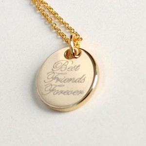 May include: Gold-colored pendant necklace with an oval charm. The charm has the words "Best Friends Forever" engraved in an elegant script. The necklace chain is also gold-colored.