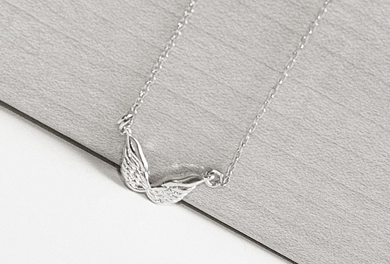 May include: A silver necklace with a delicate chain and a wing pendant. The wings are detailed and have a textured surface.