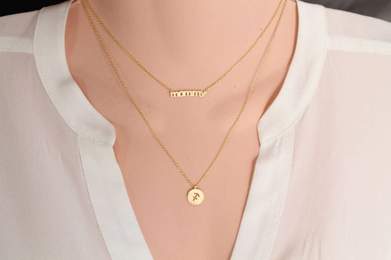 Może przedstawiać: Złoty naszyjnik z dwoma łańcuszkami. G&oacute;rny łańcuszek ma napis "mommy". Dolny łańcuszek ma zawieszkę z symbolem zodiaku Strzelca.