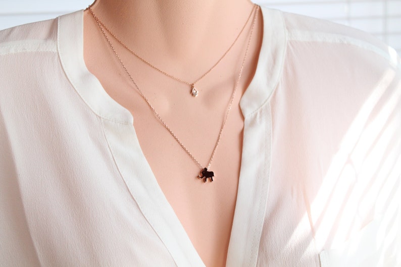 May include: A layered necklace with a small diamond pendant and a charm shaped like an elephant. The necklaces are gold-colored and worn over a white blouse.