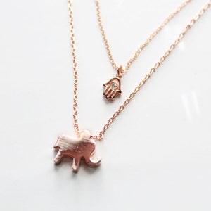 May include: A rose gold layered necklace featuring two pendants. The bottom pendant is an elephant, and the top pendant is a hand with a small gem. The necklaces are on a white background.