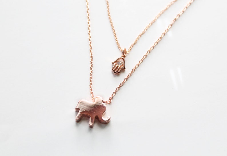 May include: A rose gold layered necklace featuring two pendants. The bottom pendant is an elephant, and the top pendant is a hand with a small gem. The necklaces are on a white background.
