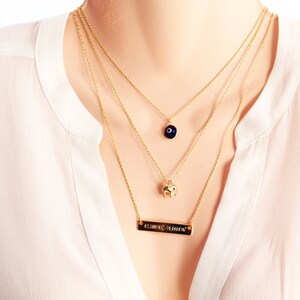 Puede incluir: Conjunto de collares dorados en capas. La cadena superior tiene un pequeño colgante de ojo azul, la cadena central tiene un colgante de elefante dorado y la cadena inferior tiene una barra rectangular grabada. Los collares se llevan sobre una blusa blanca.