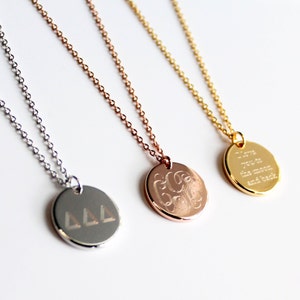 May include: Three pendant necklaces in silver, rose gold, and gold. Each necklace features a round pendant with different engraved designs: Greek letters, initials, and the phrase "I love you to the moon and back."