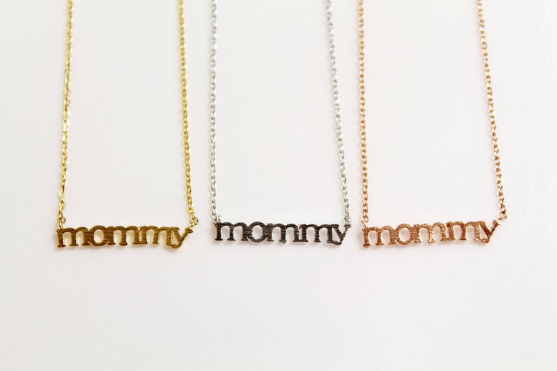 Puede incluir: Tres collares de oro con la palabra "mommy" escrita en cada uno. Los collares son de oro, plata y oro rosa.