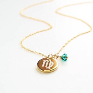 May include: A gold-colored necklace with a delicate chain. The pendant is a gold-colored disc with the Virgo symbol engraved on it. A small, teal-colored crystal bead hangs next to the pendant. The necklace is on a white background.