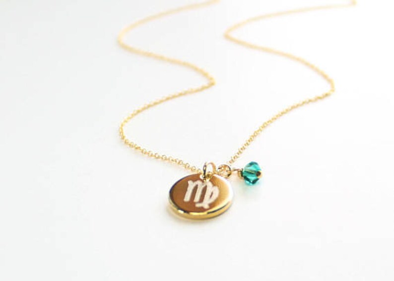 May include: A gold-colored necklace with a delicate chain. The pendant is a gold-colored disc with the Virgo symbol engraved on it. A small, teal-colored crystal bead hangs next to the pendant. The necklace is on a white background.
