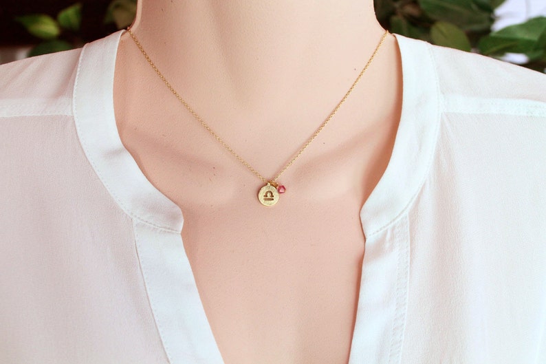 May include: Gold necklace with a Libra zodiac sign charm and a small pink gemstone.