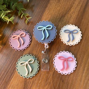 May include: A collection of five badge reels with a scalloped edge design. Each reel features a bow in a different color: pink, blue, green, and white. The reels are on a wooden surface.
