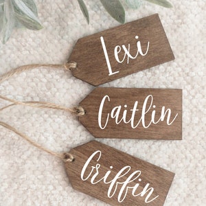 May include: Three wooden gift tags with twine, each with a different name written in white cursive: Lexi, Caitlin, and Griffin.
