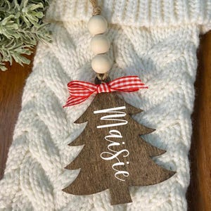 May include: A wooden Christmas tree ornament with the name "Maisie" in white script. The ornament has a red and white checkered bow and is suspended by wooden beads and twine. The background is a white cable-knit stocking.